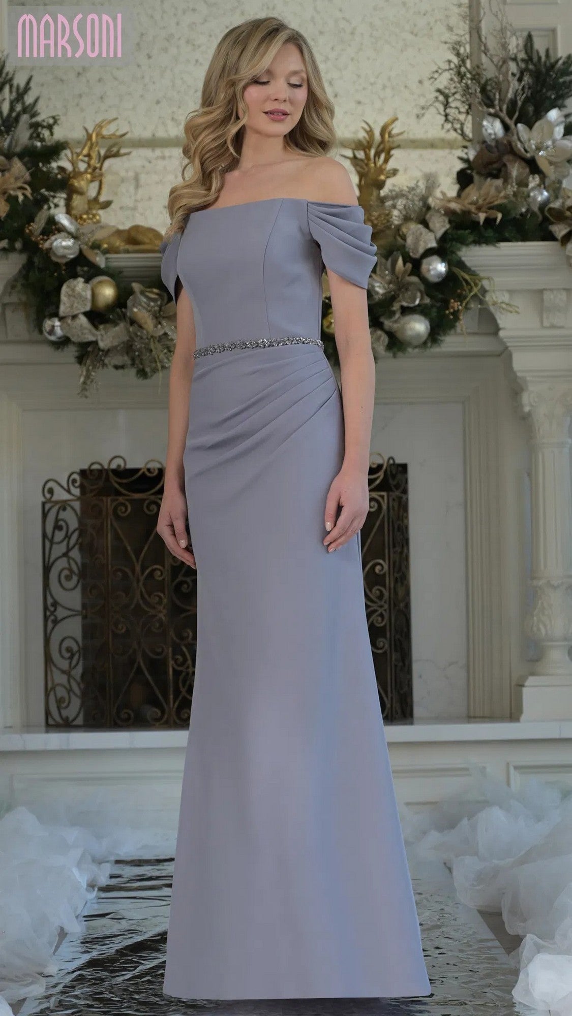 A woman stands indoors wearing the grey, floor-length Marsoni MV1352 formal dress with a beaded waistband and off-shoulder design. Festive decorations and a fireplace are in the background.